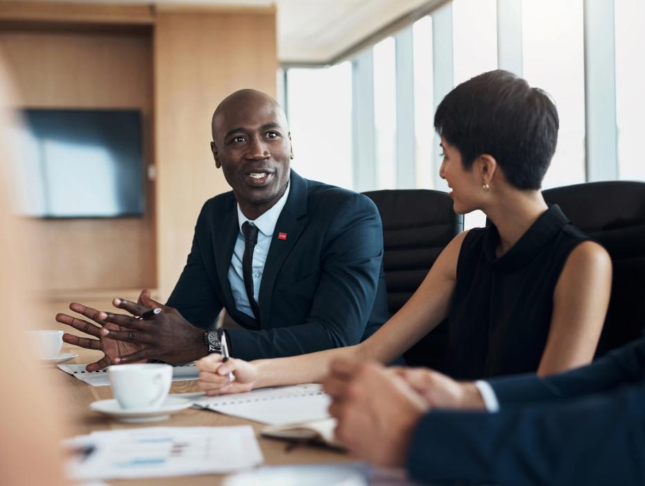 An African American man in a suit and a white woman with short hair in a dress sit at a conference table having a conversation.