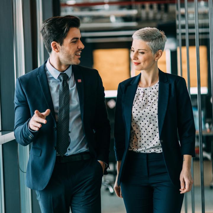 A man in suit and a woman in business wear walk down a hallway having an animated conversation.
