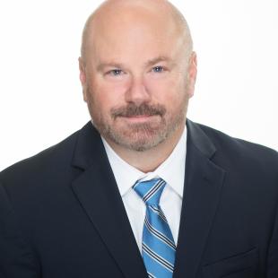 bald man with light brown beard and mustache wearing a black suit and blue striped tie.