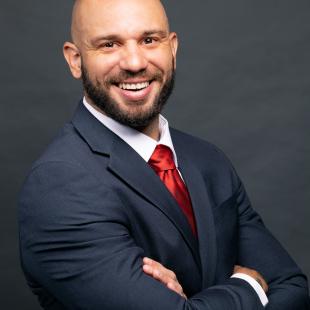 Bald olive skinned man with dark brown facial hair wearing a dark suit and red tie.