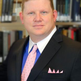 Middle aged caucasian male with light red hair in a black suit with a bright blue collared shirt