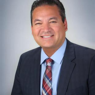 Olive skinned man in dark suit and red and blue plaid tie.